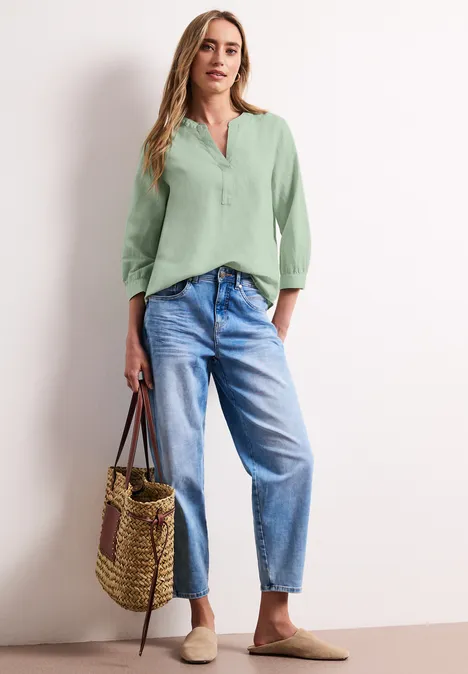Frau in hellgrüner Bluse, weitem Jeans, Strohtasche und flachen Slippern.